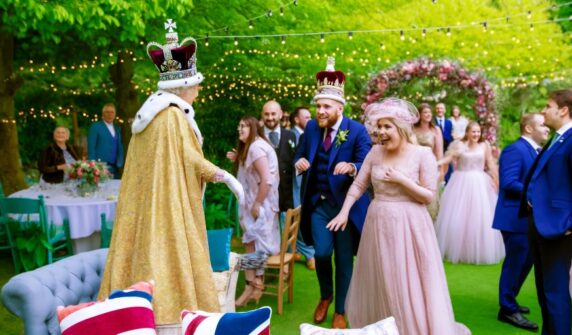 Royal Surprise: When the Queen of Great Britain accepts an invitation to a public wedding. A vibrant scene at a public wedding set in a picturesque garden. The Queen of Great Britain, dressed in regal attire with a crown, mingles with guests. The bride and groom are at the center, joyfully interacting with the Queen. The background features floral arches and fairy lights, creating a festive and celebratory atmosphere.